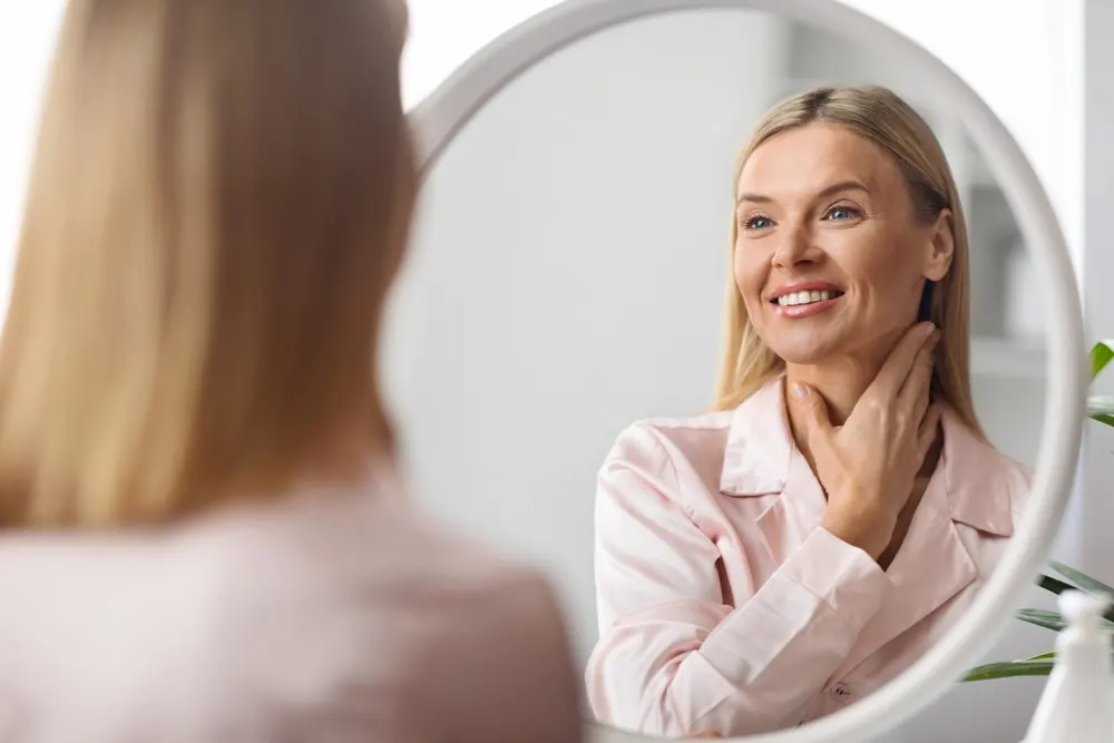 middle aged woman smiling in the mirror while examining her stunning neck and jaw